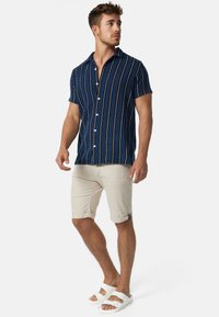 Chemise à manches courtes à rayures navy avec boutons blancs, associée à un short beige clair retroussé et des sandales blanches.