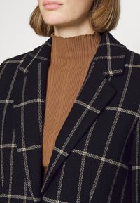 Personne portant un pull en maille côtelée marron à col haut sous un manteau noir avec des motifs à carreaux beiges et blancs, coupé au niveau du menton et des épaules.