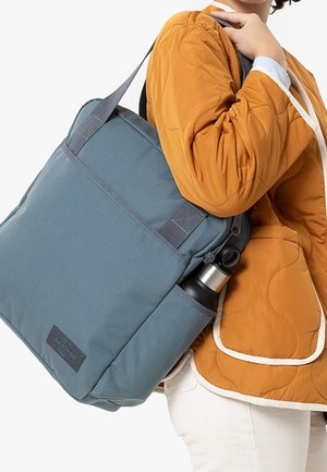 Osoba ubrana w musztardową pikowaną kurtkę, niosąca niebieską torbę na ramię z zewnętrzną kieszenią, w której trzyma czarno-srebrną butelkę na wodę.