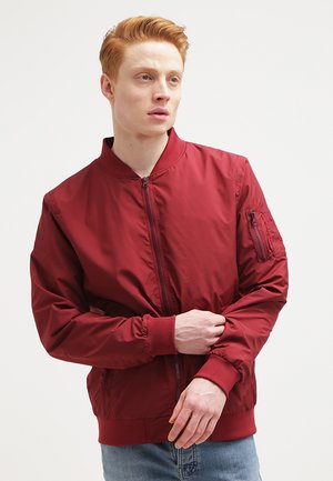 Chaqueta bomber roja hecha de material ligero, con cremallera frontal, puños acanalados y un bolsillo con cremallera en el brazo izquierdo. Textura suave.