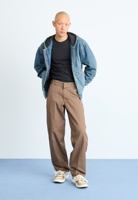 Jaqueta de ganga azul com capuz cinza, t-shirt preta, calças largas castanhas e sapatilhas claras com padrão de camuflagem. Fundo simples.