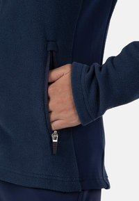 Giacca in pile blu marina presenta una tasca con zip, una consistenza morbida e polsini aderenti, mostrata con una mano poggiata all'interno della tasca.