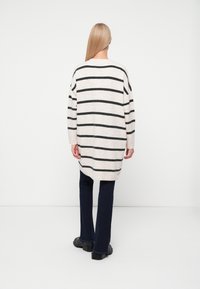 Cremfarbener und schwarzer gestreifter oversized Pullover mit langen Ärmeln, rundem Ausschnitt und weicher Textur, kombiniert mit dunklen Schlagjeans und flachen Schuhen.