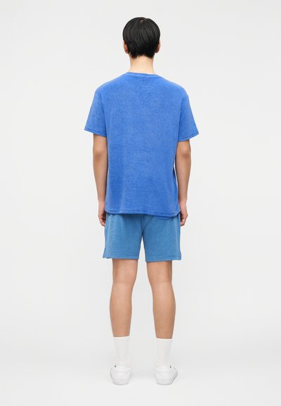 Άνδρας με την πλάτη προς το μέρος μας, φοράει μπλε κοντομάνικο πουκάμισο και αντίστοιχα μπλε σορτς, λευκές κάλτσες και λευκά αθλητικά παπούτσια, σε ένα απλό φόντο.