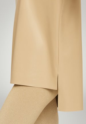 Naakte hoogglans tuniek met zijinsneden, gecombineerd met glinsterende gouden leggings. De tuniek heeft een soepele textuur, terwijl de leggings glinsteren.