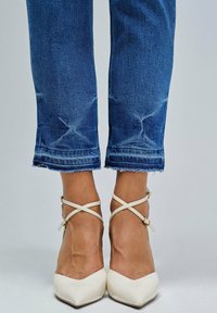 Zapatos de tacón en charol blanco con punta afilada y correas cruzadas en el tobillo, combinados con jeans desgastados de color azul medio con dobladillos deshilachados.