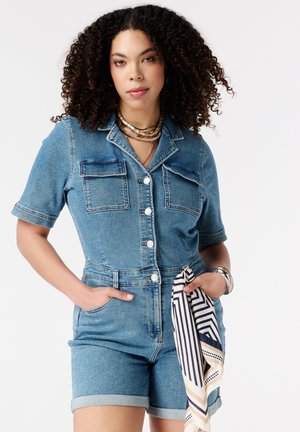 Vrouw met krullend haar die een denim korte-mouwen jumpsuit draagt, met handen in de zakken en een gestreepte sjaal die aan de lus van de riem hangt.