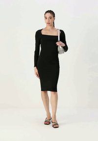 Juoda ilgomis rankovėmis prigludusi suknelė su kvadratiniu iškirpimu, iki kelių ilgio ir lygaus paviršiaus, derinama su juodais šlepetėmis ir sidabriniu rankiniu krepšiu.