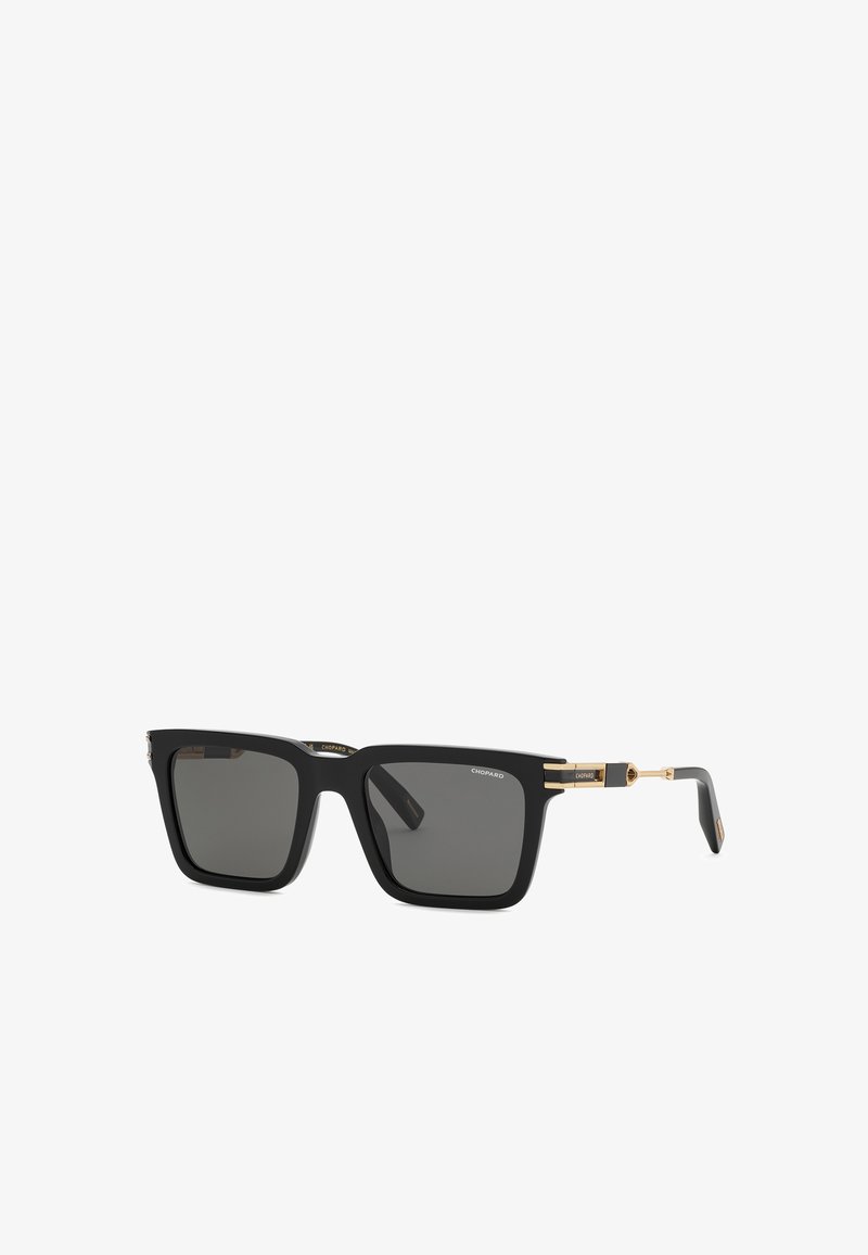 Lunettes de soleil rectangulaires noires avec des verres foncés et des accents dorés sur les branches. Nom de la marque visible dans un coin d’un verre.