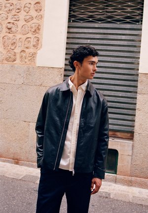Young man with dark curly hair wearing a black leather jacket and white shirt stands on a street, looking to the side.