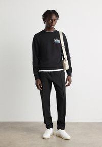 Zwarte sweatshirt met een grafische patch, gecombineerd met zwarte broeken en witte sneakers. De outfit wordt aangevuld met een schoudertas in beige.