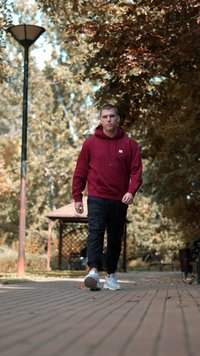 Moški hodi po tlakovani poti, oblečen v burgundski kapuco, črne cargo hlače in bele superge, obdan z jesenskim listjem.