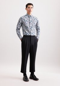 Uomo con una camicia floreale a maniche lunghe e bottoni, infilata in pantaloni neri a vita alta con risvolti e scarpe da eleganza nere, in piedi.