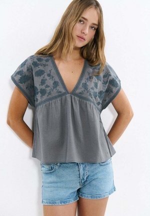 Jeune femme portant un chemisier ample gris brodé de fleurs avec un profond col en V et un short en jean bleu clair, debout contre un mur blanc.