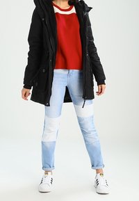 Svart huva parka med en fluffig foder, kombinerad med en röd tröja och ljusblå färgblockerade jeans. Vita sneakers med svarta detaljer.