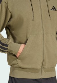 Sweat à capuche zippé vert olive en tissu doux, avec des accents noirs à trois bandes sur les manches et les poches avant.