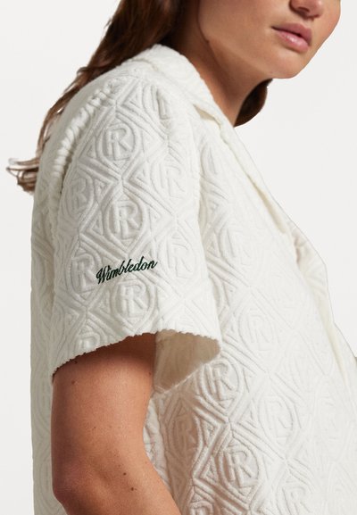 Chemise blanche à manches courtes avec un motif géométrique texturé et un détail brodé "Wimbledon" en vert sur le poignet.