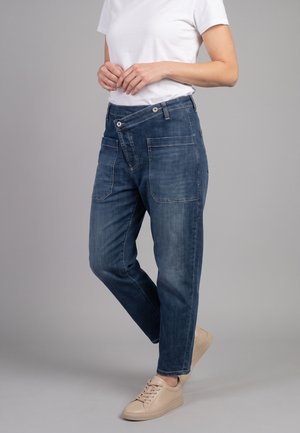 Persona que lleva vaqueros azules con bragueta de botones superpuestos y camiseta blanca, de pie sobre un pie con zapatillas beige sobre fondo gris.