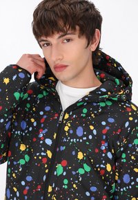 Jachetă neagră cu un model colorat de stropi de vopsea; are un închidere cu fermoar și o glugă ajustabilă; textură netedă, material sintetic.