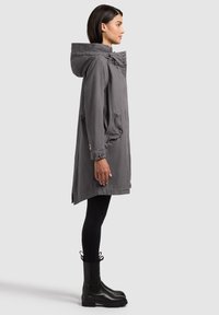 Donna indossa un cappotto grigio con cappuccio di media lunghezza, leggings neri e stivali neri fino a metà polpaccio, in piedi di profilo su uno sfondo uniforme.