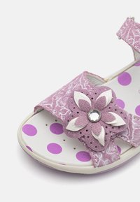 Lila Stoffsandale mit floraler Akzentuierung, weißen synthetischen Blütendetails und gepunktetem Innenlauf. Mit verstellbarem Riemen.
