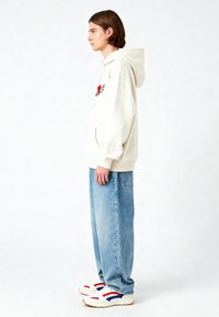 Felpa leggera beige con tasca frontale e design a forma di cuore rosso; indossata con jeans in denim blu strappati e sneakers bianche con dettagli rossi e blu.