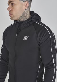Schwarze Zip-Hoodie aus glattem Stoff, mit hohem Kragen, weißen Akzenten und einem kleinen gestickten Logo auf der Brust.