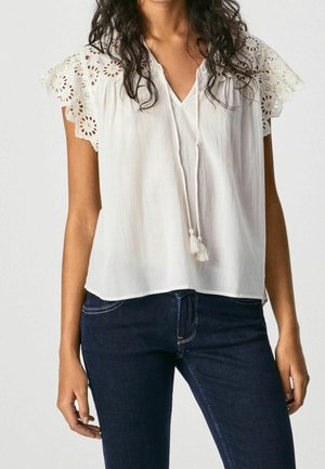 Mujer que lleva una blusa blanca sin mangas con hombros de encaje y lazos con borlas, combinada con vaqueros azul oscuro.