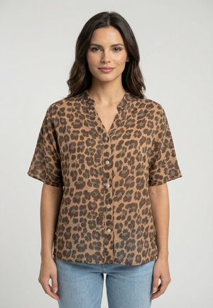 Vrouw met donker haar draagt een bruin overhemd met korte mouwen in luipaardprint en een lichtblauwe spijkerbroek, staand tegen een effen lichte achtergrond.