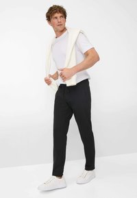 Witte t-shirt, zwarte broek met een trekkoord en witte sneakers. De outfit is casual en aangesloten, met een crèmekleurige trui over de schouders gedrapeerd.