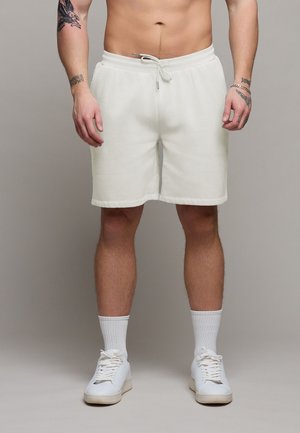 Mann iført hvite shorts med snøring, hvite crew-sokker og hvite joggesko, stående foran en ensfarget grå bakgrunn.