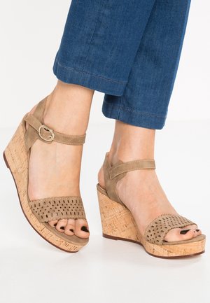 Beżowe sandały na koturnie z perforowanymi zamszowymi paskami, z klamrą w okolicy kostki i teksturowanymi platformami z korka. Otwarty nos ukazujący pomalowane paznokcie.