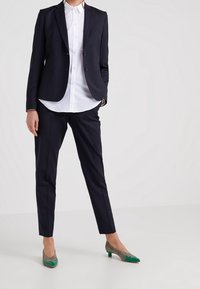 Giacca blu navy, camicia bianca, pantaloni scuri su misura e scarpe con tacco basso con un design grigio e verde. Tessuto morbido e vestibilità strutturata.
