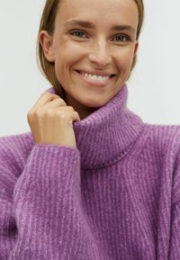 Pulover purpuriu cu dungi, cu guler înalt și un model texturat, realizat dintr-un material moale. Modelul zâmbește în timp ce își ține gulerul.