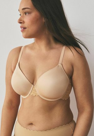 Femeie purtând un sutien nude cu burete și detalii din dantelă, împreună cu chiloți cu talie înaltă asortați, privind spre stânga pe un fundal neutru.