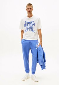 Weißes T-Shirt mit blauer Aufschrift "TOMMY ATH.DPT JEANS NEW YORK", kombiniert mit blauen Jogginghosen und einem blauen Sweatshirt. Weiße Sneakers.