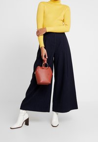 Maglione giallo a coste con collo alto, pantaloni ampi a righe navy, stivaletti bianchi alla caviglia e una piccola borsa esagonale rossa con manico ad anello.