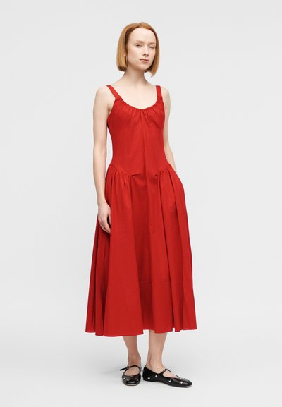 Κόκκινο βαμβακερό φόρεμα με εφαρμοστή μπούστα, σούρες στη λαιμόκοψη και φαρδιά φούστα. Φοριέται με μαύρα φλατ παπούτσια με διακοσμητικές λεπτομέρειες.