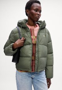 Oliven grønn puffjakke med hette, med glidelås foran, quiltet tekstur og en liten logo-detalj. Kombinert med en mønstret genser og jeans.
