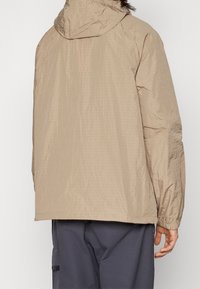 Beige huva jacka med ett texturerat, lättviktig material. Har elastiska ärmslut och en avslappnad passform, vilket ger komfort och rörelsefrihet.