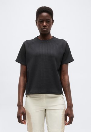 Lühikeste juustega naine, kes kannab musta tekstuurse lühikese varrukaga särki ja heledaid beežikaid lühikesi pükse, seisab ühevärvilise heleda tausta ees.