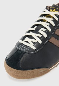 Sneaker in pelle nera con lacci crema, dettagli in suede marrone e tre strisce marroni. Suola in gomma texturizzata con una punta liscia.
