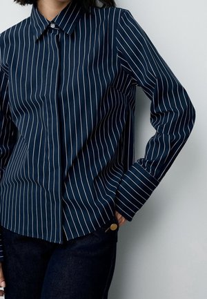 Femme portant une chemise bleu marine à manches longues avec des rayures verticales blanches et un jean foncé, main dans la poche, sur un fond gris clair.