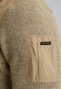 Beige gebreide trui met textuurstof en een patchzak van een glad, tan materiaal met een zwart label en gele tekst.