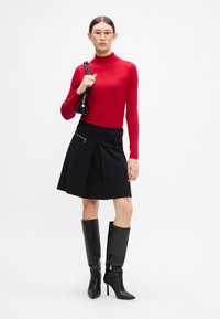 Czerwona sweter turtleneck, czarna spódnica plisowana z detalem zamka, długie czarne skórzane kozaki za kolano, trzymająca małą czarną torebkę.