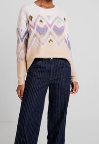Multicolored sweater with a diamond pattern in beige, purple, and gold. Made of soft material with sequins and ribbed cuffs. Paired with navy striped pants.