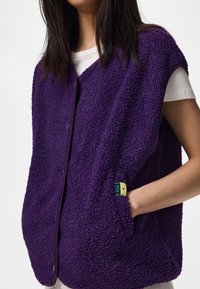 Γυναίκα που φοράει μοβ fleece αμάνικο γιλέκο με πλευρικές τσέπες πάνω από ένα λευκό κοντομάνικο πουκάμισο, χέρι στην τσέπη του γιλέκου.