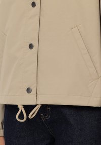 Béžová bunda s hladkou texturou, stříbrnými patentkami a nastavitelným spodním lemem se šňůrkou. Má boční kapsu a minimalistický design.