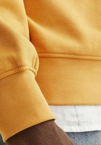 Tessuto di felpa gialla con maglia testurizzata, dotato di polsini a coste e cucitura della spalla liscia, indossato sopra una maglietta bianca.