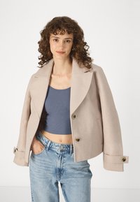 Μπεζ ζακέτα από μίγμα μαλλιού με φαρδύ γιακά και δύο κουμπιά. Συνδυασμένη με ένα γκρι εφαρμοστό τοπ και ανοιχτό μπλε ψηλόμεσα τζιν.